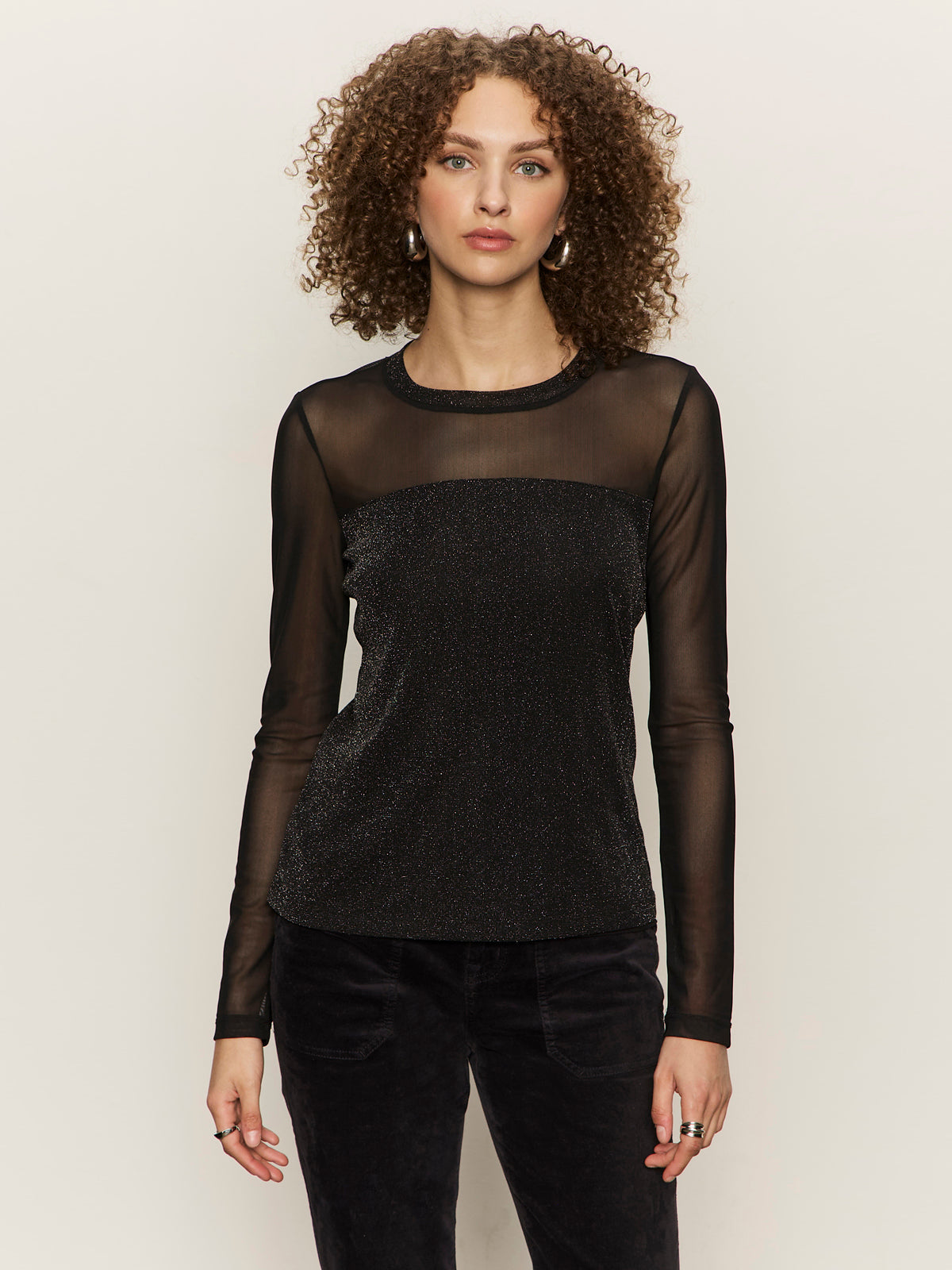 A woman with curly hair wears the Sanctuary Clothing sparkle mesh crew tee black and black pants, standing against a plain light background, looking directly at the camera.
