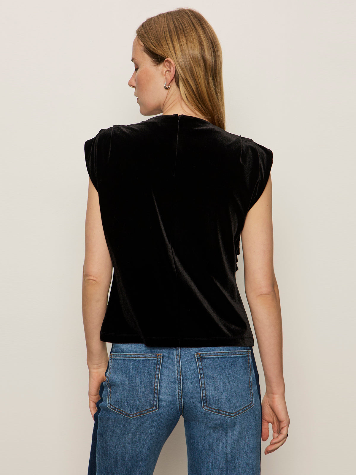 A woman with straight, light brown hair is shown from the back wearing Sanctuary Clothing's pleated sleeveless top in black and blue jeans with visible back pockets, standing before a plain light-gray background.