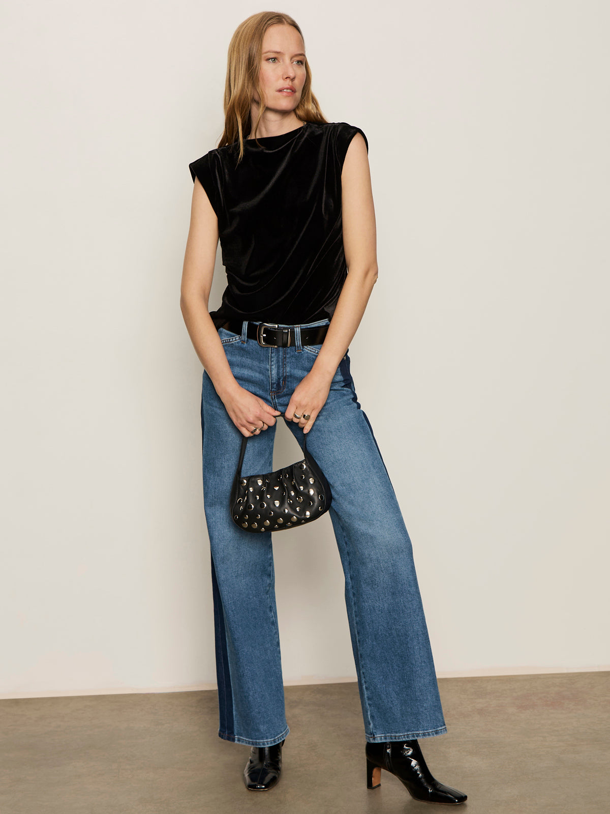 A woman stands against a plain wall wearing the Sanctuary Clothing pleated sleeveless top in black, wide-leg blue jeans, black heeled boots, and holding a small black handbag with a silver pattern.