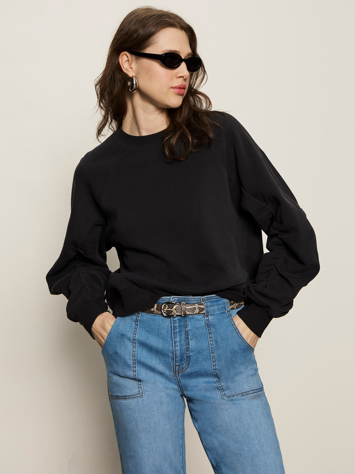 A woman wearing the Sanctuary Clothing shirred sleeve sweatshirt in black and high-waisted blue jeans, with black sunglasses and her hands in her pockets, stands against a plain beige background.