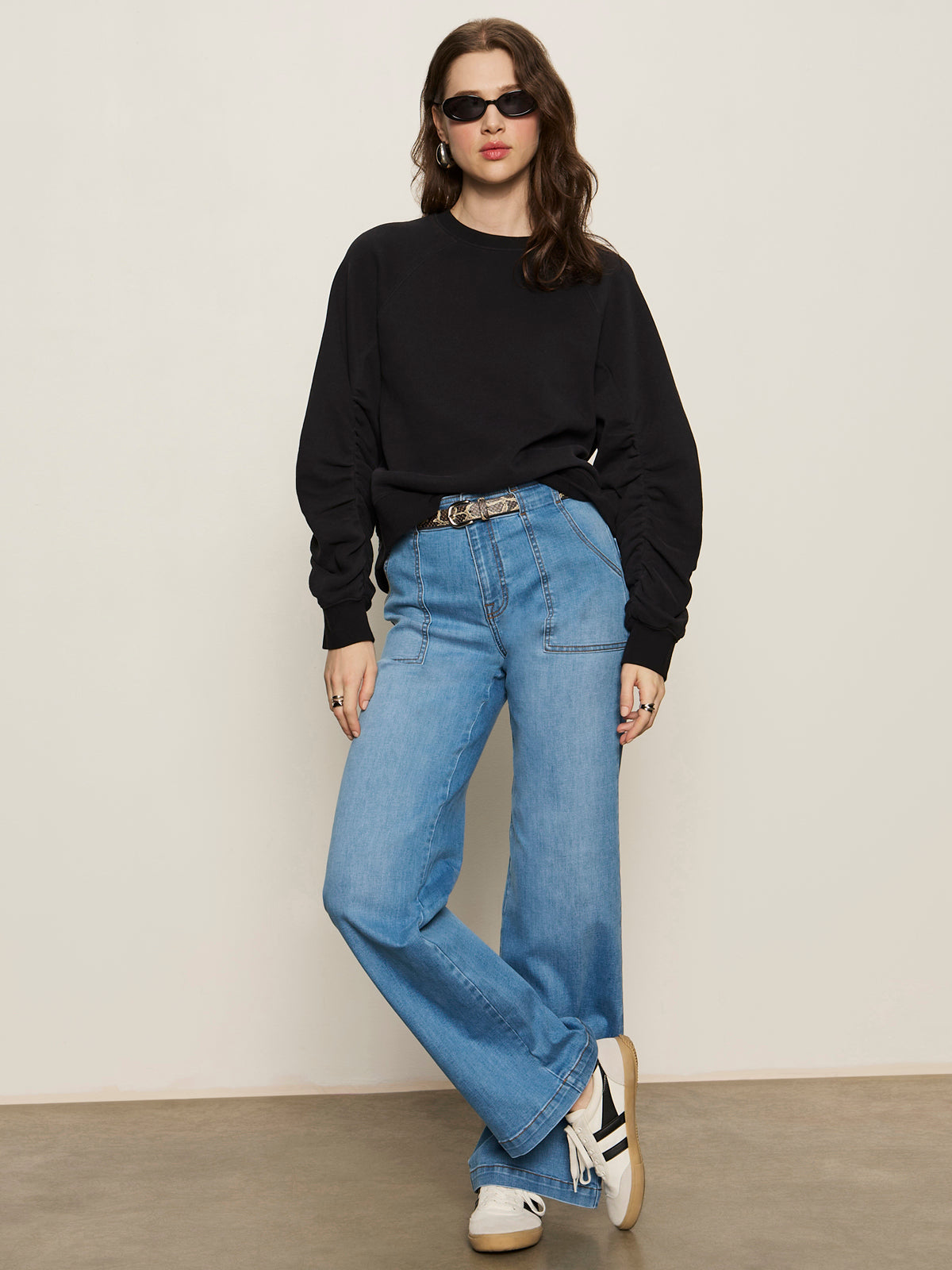 A woman with wavy brown hair stands against a plain background wearing a Sanctuary Clothing shirred sleeve sweatshirt in black, light blue wide-leg jeans, white sneakers, sunglasses, a belt, and has her hands in her pockets.
