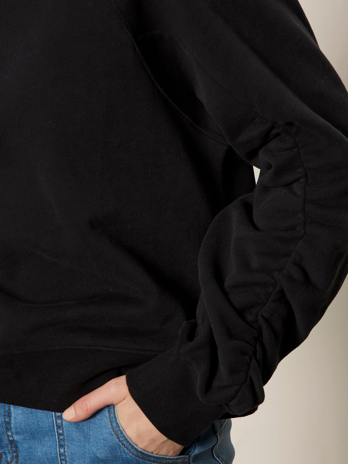 A person wears the Sanctuary Clothing shirred sleeve sweatshirt in black, hand on the hip of blue jeans, shown from the side against a neutral background.