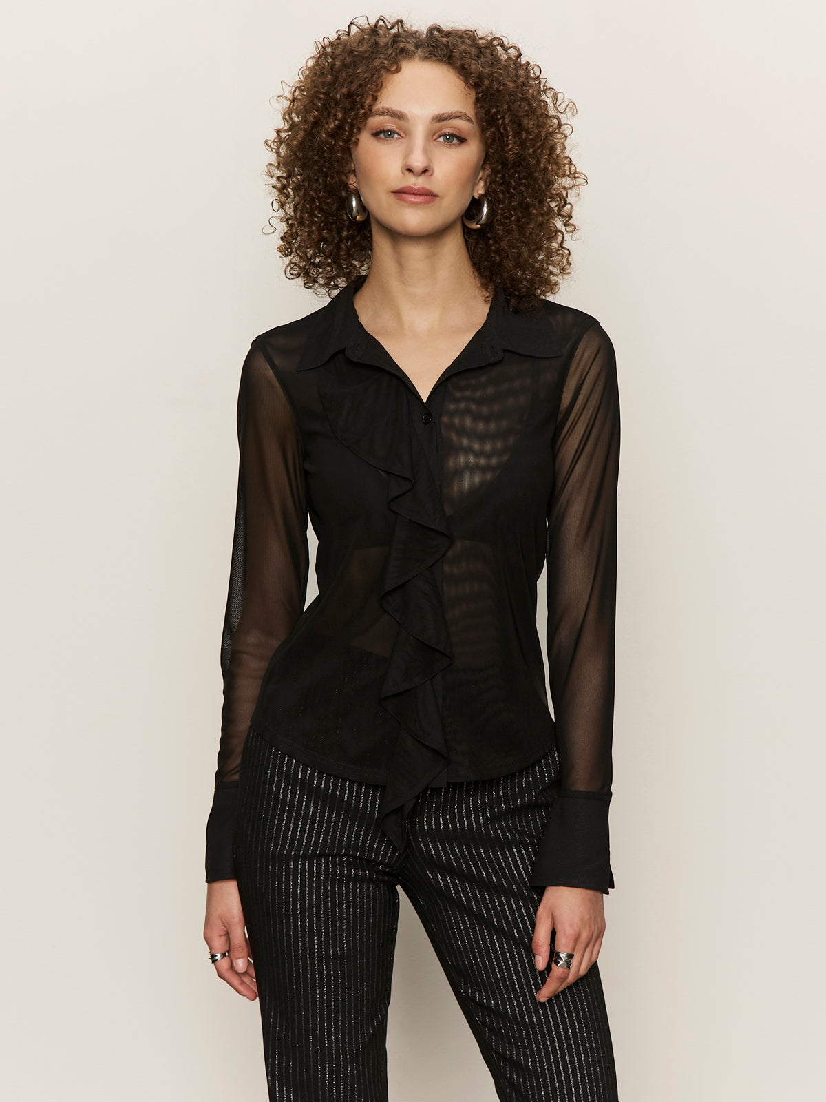 A woman with curly hair wears the Sanctuary Clothing dreamland ruffle top in black and pinstriped black pants, standing against a plain light background.