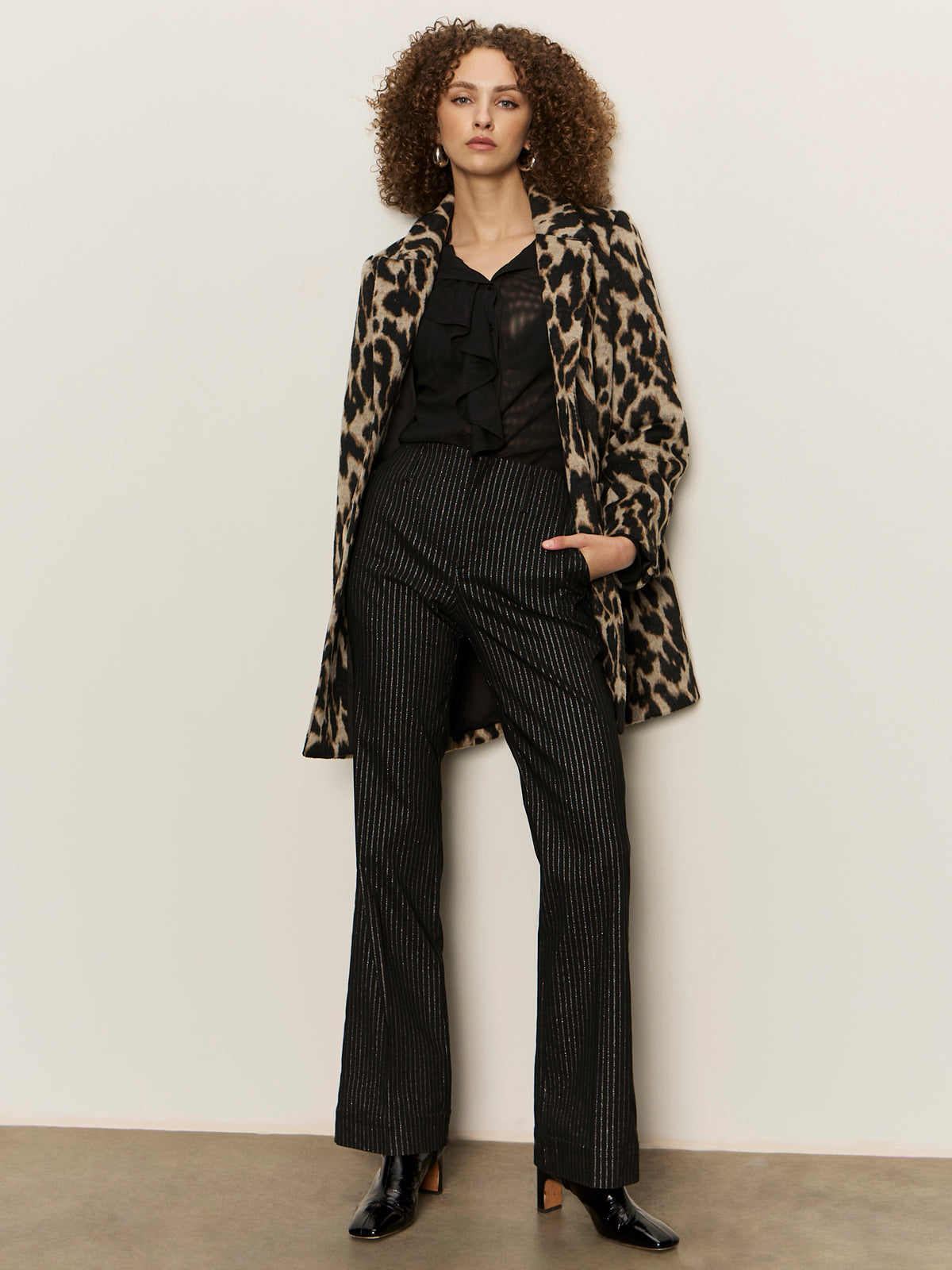 A person stands against a beige wall wearing Sanctuary Clothing„¢s dreamland ruffle top in black, paired with a leopard print coat, black pinstriped trousers, and black heeled boots. They have curly hair and a neutral expression.
