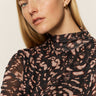 A woman with fair skin and straight blonde hair models the Sanctuary Clothing long sleeve mesh mock top nocturnal, featuring a black and pink pattern, along with small gold hoops against a plain light background.