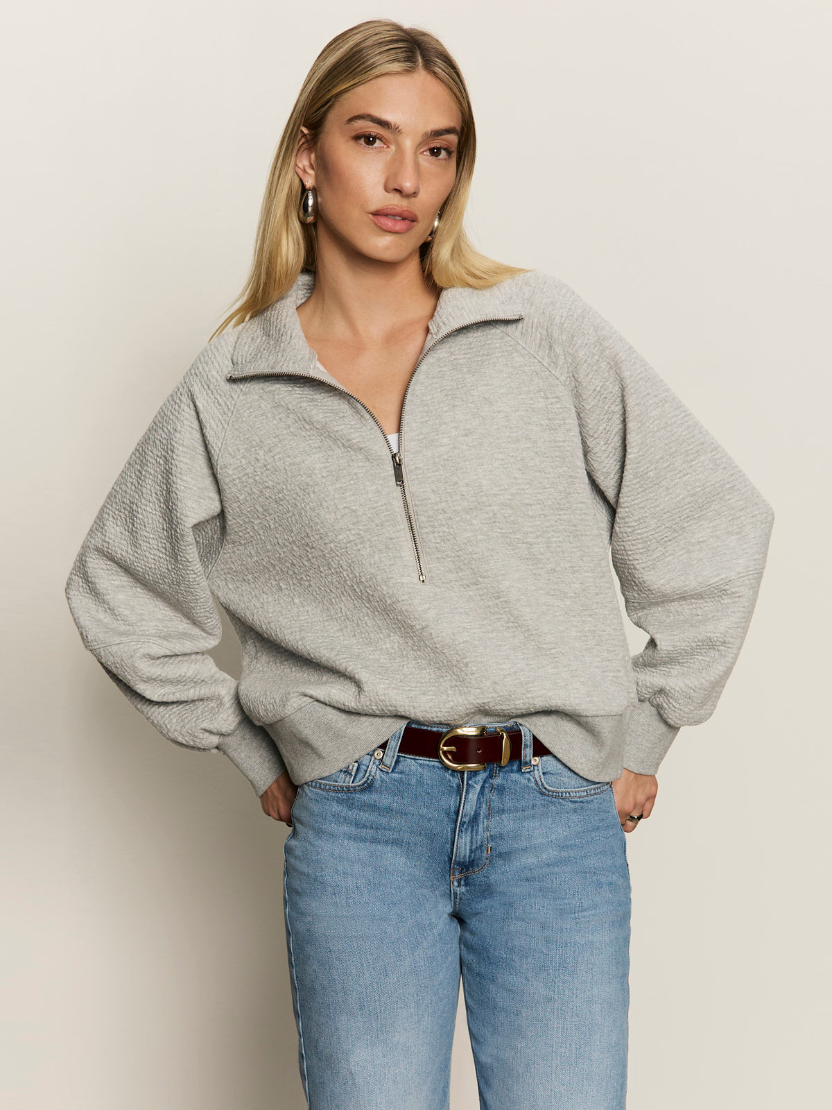 A woman with long blonde hair wears the Sanctuary Clothing textured zip up popover in heather grey and blue jeans, standing with one hand in her pocket against a plain, light background.