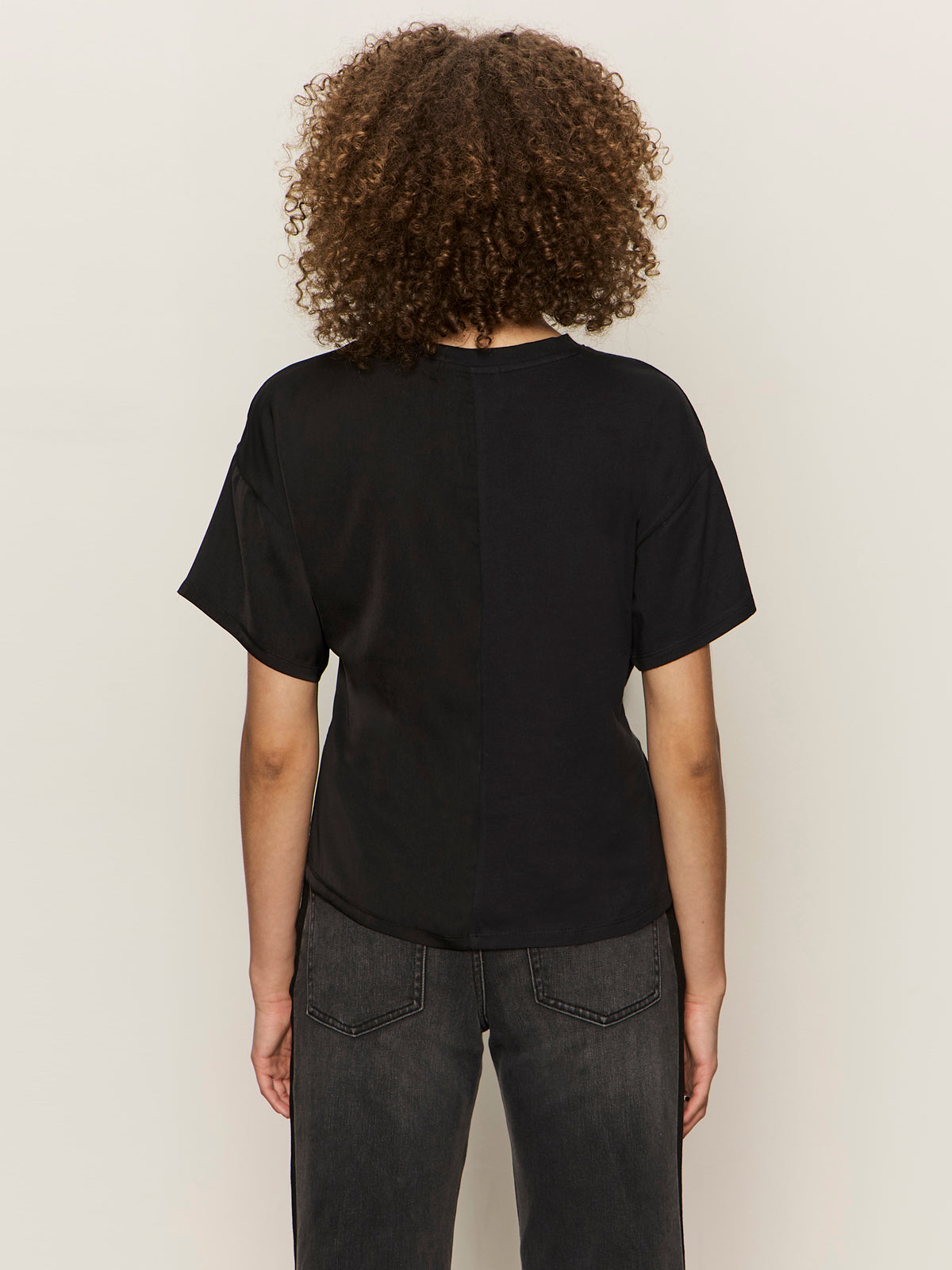 A person with curly hair faces away from the camera, wearing the Sanctuary Clothing satin mix tee in black and dark jeans against a plain light background.