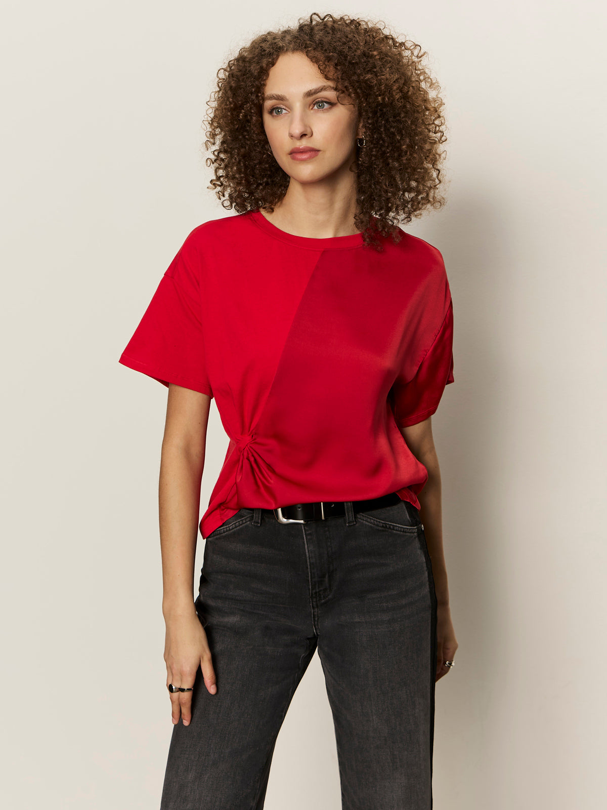 A woman with curly hair wears the Sanctuary Clothing satin mix tee in cherry lacquer, styled with a side knot and paired with black jeans, standing against a light background and looking slightly to her left.