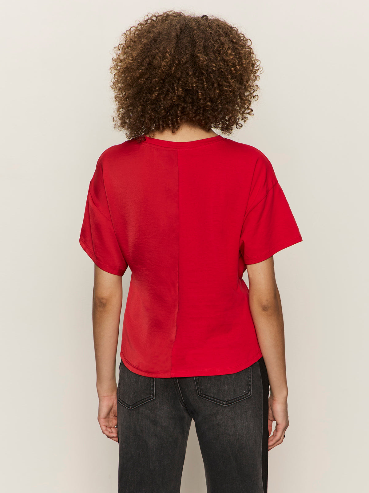 A person with curly hair faces away from the camera, wearing Sanctuary Clothing's satin mix tee in cherry lacquer and dark jeans against a plain, light background.