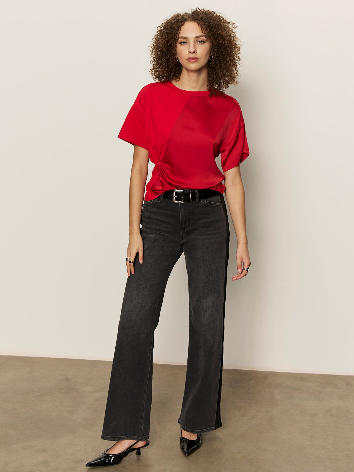 A woman with curly hair wears the Sanctuary Clothing satin mix tee in cherry lacquer, paired with black belted jeans and black heels. She stands on a smooth floor against a plain, light wall, looking relaxed.