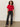 A woman with curly hair wears the Sanctuary Clothing satin mix tee in cherry lacquer, paired with black belted jeans and black heels. She stands on a smooth floor against a plain, light wall, looking relaxed.