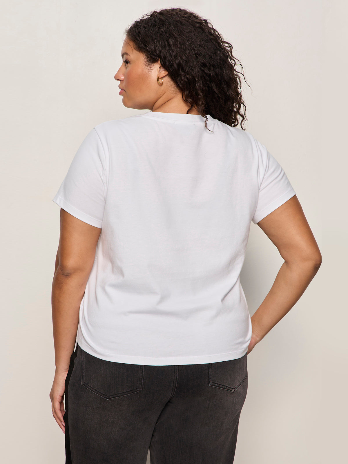 A woman with curly hair, wearing Sanctuary Clothing's embellished tee in white (extended sizing) and dark jeans, stands with her back to the camera against a neutral background.