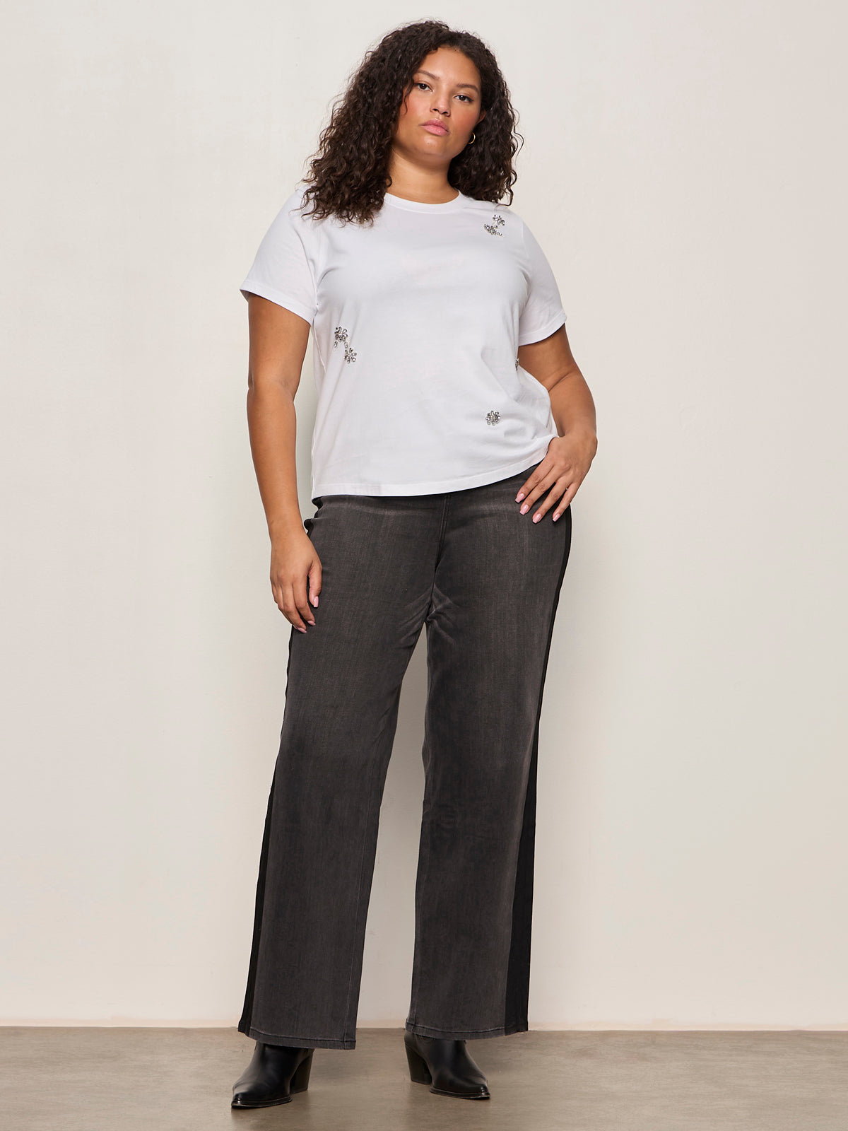 A woman with curly hair wears the Sanctuary Clothing embellished tee white extended sizing, paired with dark wide-leg jeans and black ankle boots, standing against a plain light-colored wall.