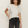 A woman with curly hair wears the Sanctuary Clothing embellished tee in white, paired with black jeans and a black belt, standing against a plain light background.