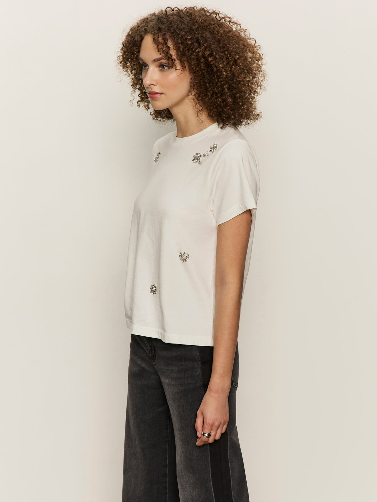 A person with curly hair stands sideways against an off-white background, wearing the Sanctuary Clothing embellished tee white and dark wide-leg jeans.