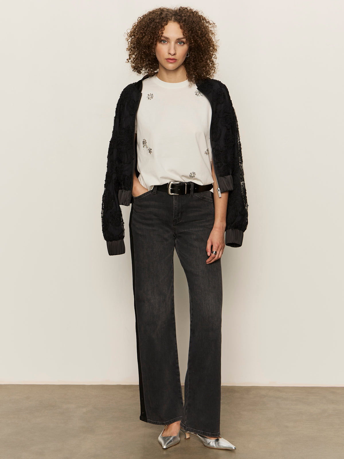 A person with curly hair stands against a plain background, wearing the Sanctuary Clothing embellished tee in white, paired with a black textured jacket, black wide-leg jeans, a black belt, and silver pointed-toe shoes.