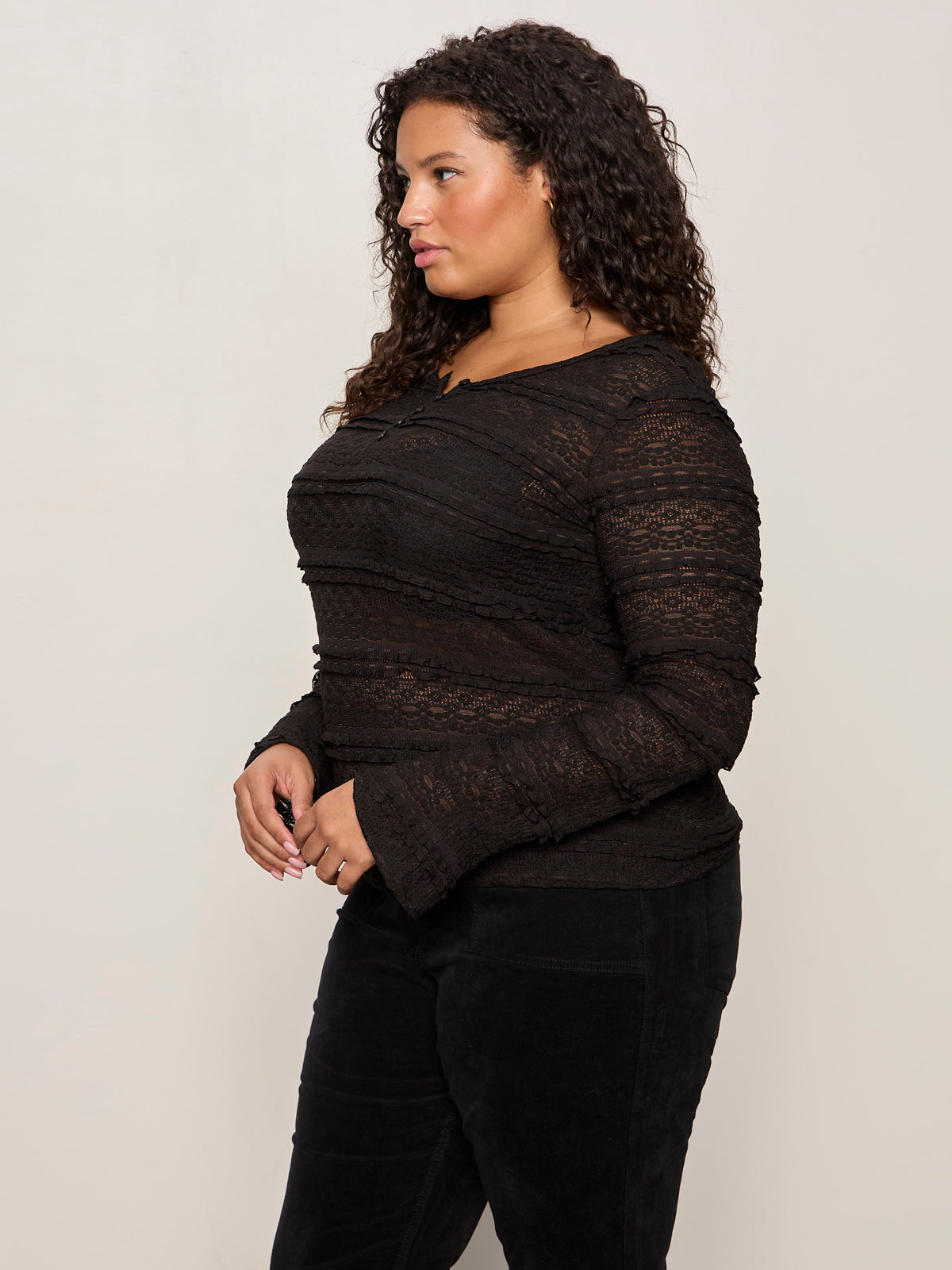 A woman with curly hair wears the Sanctuary Clothing lovely lace button front top in black (extended sizing) and black pants, standing in profile against a plain light background.