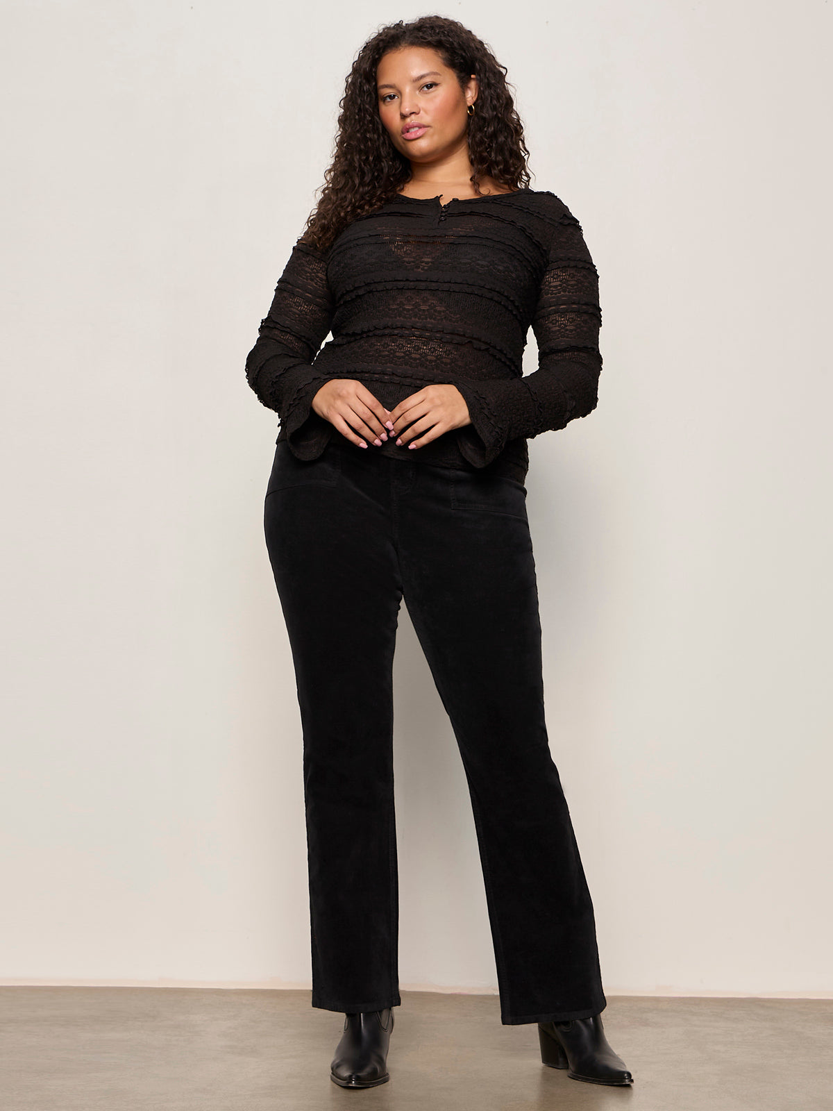 A woman with curly hair and a confident look stands against a plain wall, wearing the Sanctuary Clothing "lovely lace button front top black extended sizing," paired with black pants and ankle boots. One hand rests relaxed by her side.