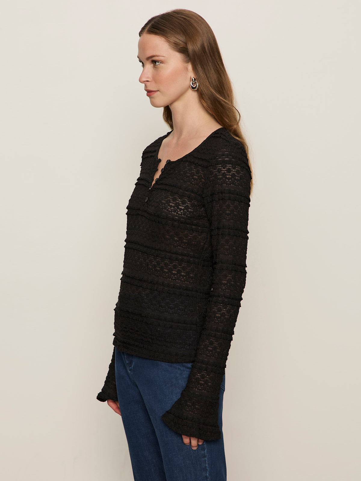 A woman with long brown hair wears the Sanctuary Clothing lovely lace button front top in black and blue jeans, standing sideways facing left against a plain light-colored background.