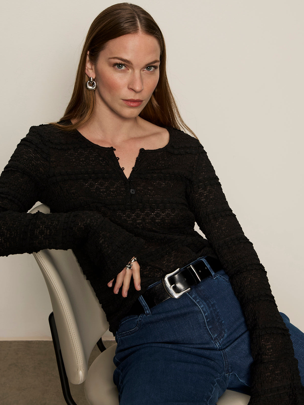 A woman with long brown hair sits on a white chair, wearing Sanctuary Clothing's lovely lace button front top in black, paired with blue jeans, a black belt, and silver earrings. She looks confidently at the camera against a plain background.