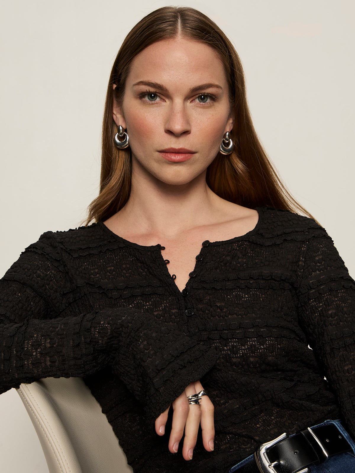 A woman with long brown hair wears the Sanctuary Clothing lovely lace button front top in black and silver hoop earrings, sitting on a light-colored chair. She looks at the camera with a neutral expression, her hand showing silver rings.