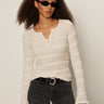 A woman with curly hair wears black sunglasses, black jeans, and a black belt. She stands against a plain light background in the Sanctuary Clothing lovely lace button front top in vintage ivory.