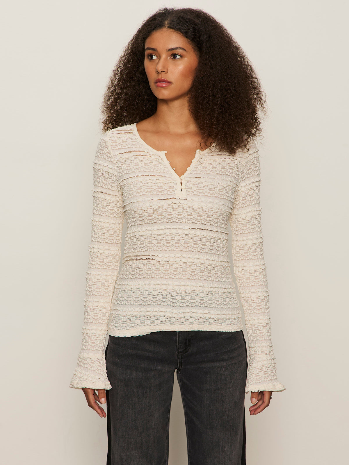 A woman with curly hair wears the Sanctuary Clothing lovely lace button front top in vintage ivory and black jeans, standing against a plain light background.