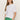 A woman with long light brown hair wears clear glasses and green pants, styling the Sanctuary Clothing boatneck ribbed top in white, standing against a light background with one hand in her pocket.