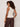A woman with curly hair wears the Sanctuary Clothing “friday rib tank collagen” sleeveless cream top with brown pants, standing against a light wall and facing away from the camera, accessorized with bangles on her wrist.