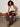 A woman with curly hair sits on a modern bench in Sanctuary Clothing’s friday rib tank collagen, styled with loose brown pants, sandals, and simple jewelry. The setting is minimal and neutral.