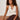 A woman with curly hair sits on a bench, thoughtfully gazing to the side. She wears the Sanctuary Clothing friday rib tank collagen in cream, brown pants, and accessorizes with two chunky bracelets.