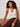 A woman with curly hair sits on a bench, thoughtfully gazing to the side. She wears the Sanctuary Clothing friday rib tank collagen in cream, brown pants, and accessorizes with two chunky bracelets.