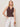 A blonde woman wears the Sanctuary Clothing friday rib tank in rich soil with white lace-up pants, accessorized with bangles and a necklace. She poses with her left hand in her pocket against a light neutral background.