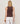 A person with shoulder-length blonde hair is seen from behind, wearing the Sanctuary Clothing Friday Rib Tank in Rich Soil and white pants. They have a chunky brown bracelet on their left wrist, standing against a plain light background.
