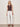 A woman stands against a plain light wall, wearing the Sanctuary Clothing friday rib tank in rich soil, white wide-leg pants with lace-up front detail, brown sandals, and gold and brown bangles.