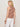 A person with shoulder-length blonde hair wears the Sanctuary Clothing campari tank in clay stripe and white pants, accessorized with bracelets on their right wrist. They stand facing away against a plain light background.