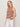 A woman with blonde hair models the Sanctuary Clothing campari tank clay stripe, a sleeveless brown and white striped top, paired with white pants. She stands against a plain light background, gazing at the camera with a relaxed expression.