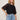A woman with long, wavy blonde hair stands against a plain background, confidently modeling the Sanctuary Clothing crochet sleeve belle tee in black with lacy short sleeves and light blue jeans, one hand resting on her hip.