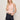 A woman with blonde hair wears the sprinter rib tank in bleached pink by Sanctuary Clothing and dark denim shorts with a tan belt. She stands against a plain light background, one hand in her pocket and the other touching her hair.
