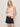 A woman with blonde hair wears the sprinter rib tank in bleached pink by Sanctuary Clothing and dark denim shorts with a tan belt. She stands against a plain light background, one hand in her pocket and the other touching her hair.