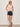 A woman wears Sanctuary Clothing's Sprinter Rib Tank in Bleached Pink, paired with dark denim shorts, a belt, black sandals, and sunglasses as she stands against a plain light wall with her hands in her pockets.