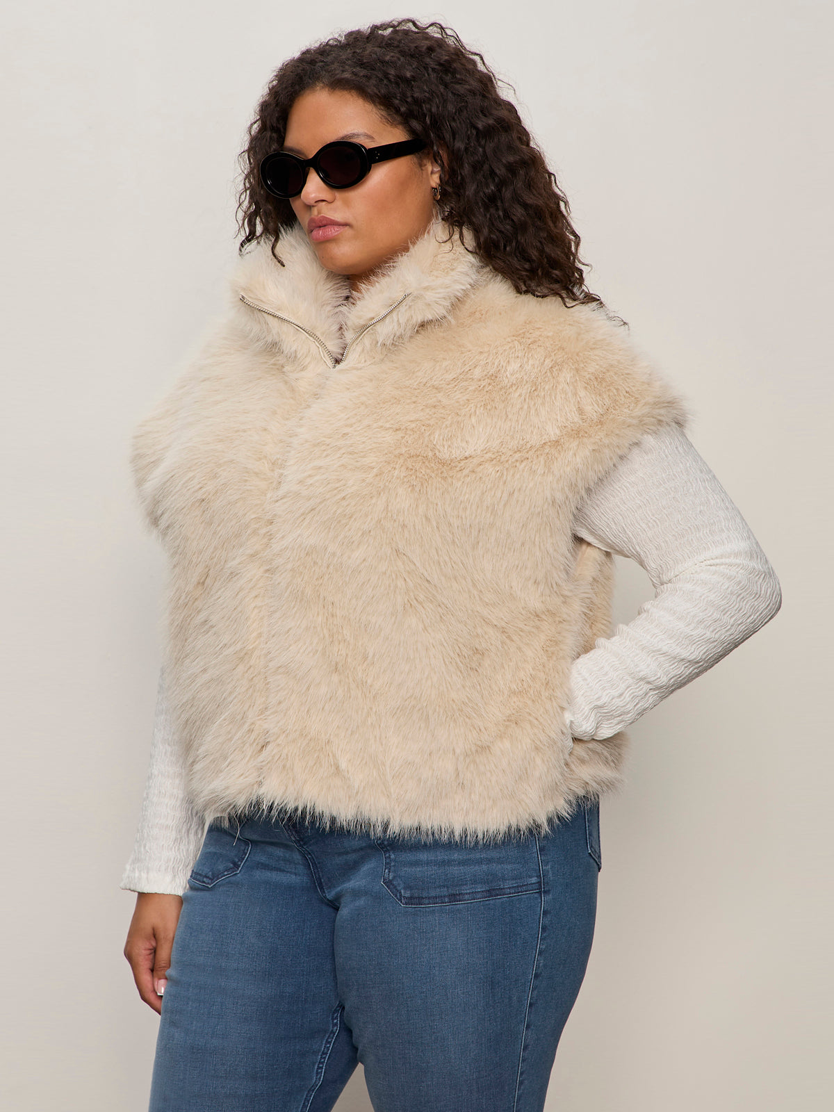 A woman in Sanctuary Clothing's silky fur vest in almond cream (extended sizing) over a ribbed white long-sleeve top, blue jeans, and dark sunglasses stands against a plain light background.