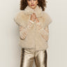 A woman with curly hair wears the silky fur vest in almond cream by Sanctuary Clothing over a lace top and metallic gold pants, standing against a plain light background.