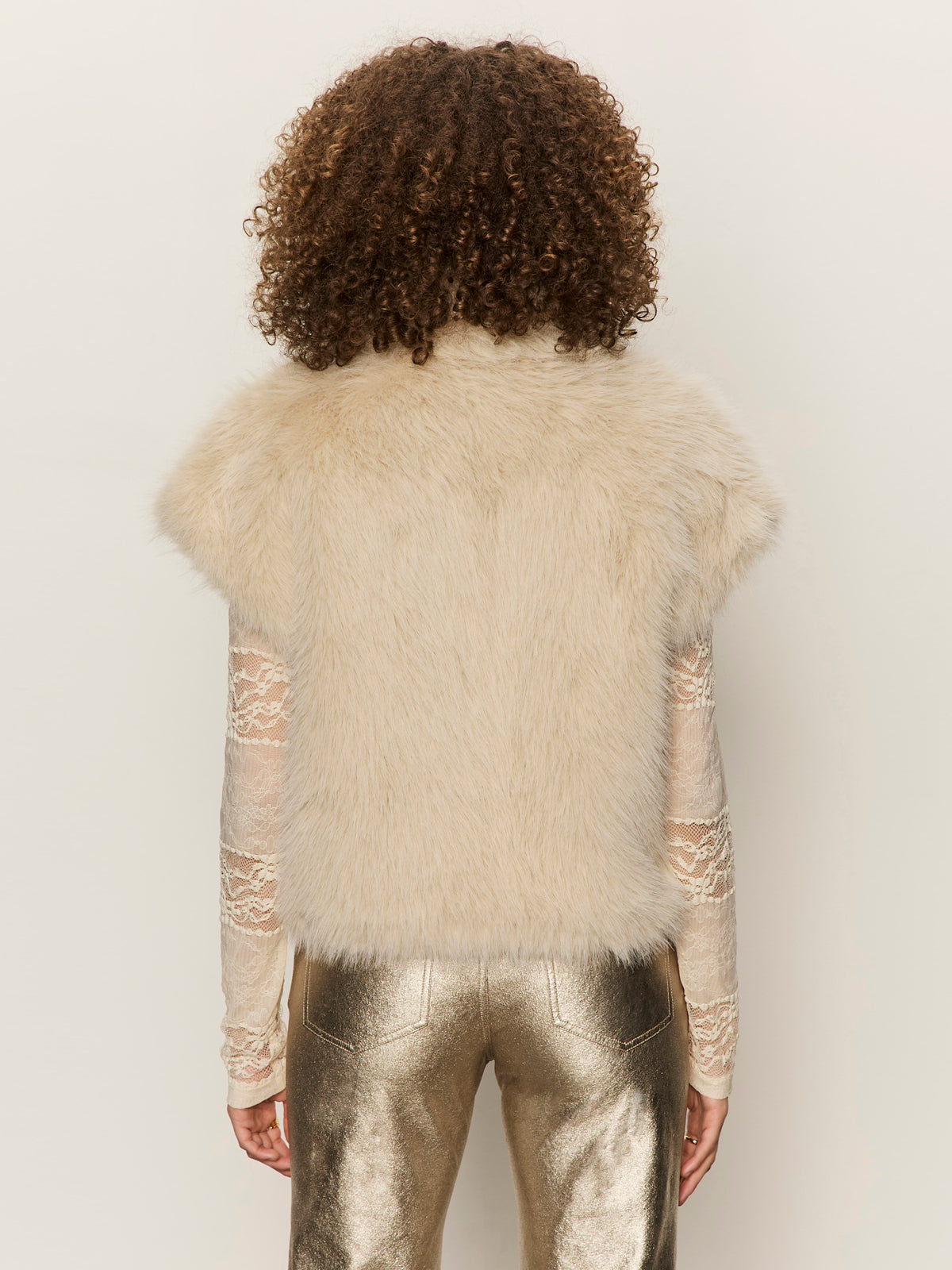A person with curly hair stands facing away, wearing the Sanctuary Clothing silky fur vest in almond cream petite sizing over a long-sleeve lace top and metallic gold pants, against a plain light background.