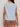 A woman is seen from the back wearing Sanctuary Clothing’s easy sweater tee in blue aura (petite sizing) and high-waisted light brown shorts with back pockets, standing against a plain, light background.