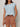 A woman wears the Sanctuary Clothing easy sweater tee in blue aura (petite sizing), tucked into high-waisted, rolled brown shorts with a dark brown belt, standing against a plain light background.