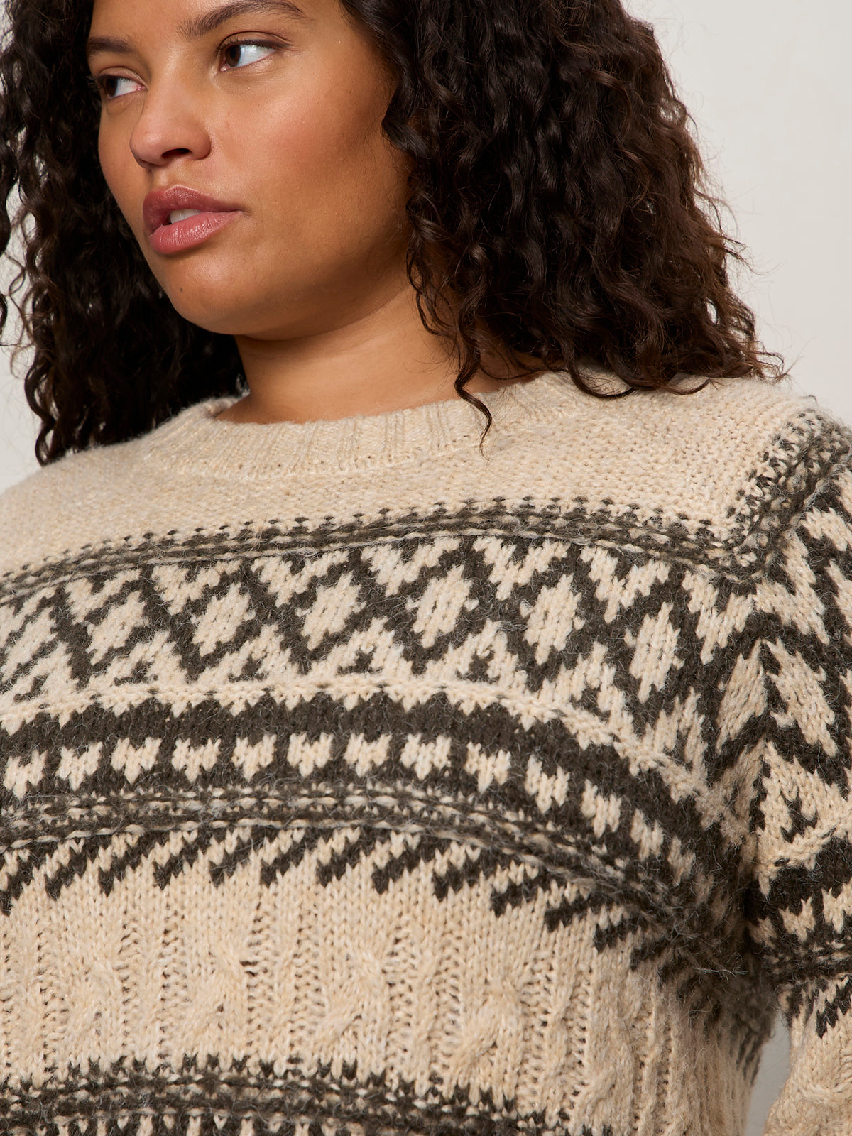 A person with curly dark hair models the Sanctuary Clothing fairisle sweater in light oat multi (extended sizing), showing its black and gray geometric chest and shoulder pattern. The image is cropped to highlight the upper sweater and partial face.