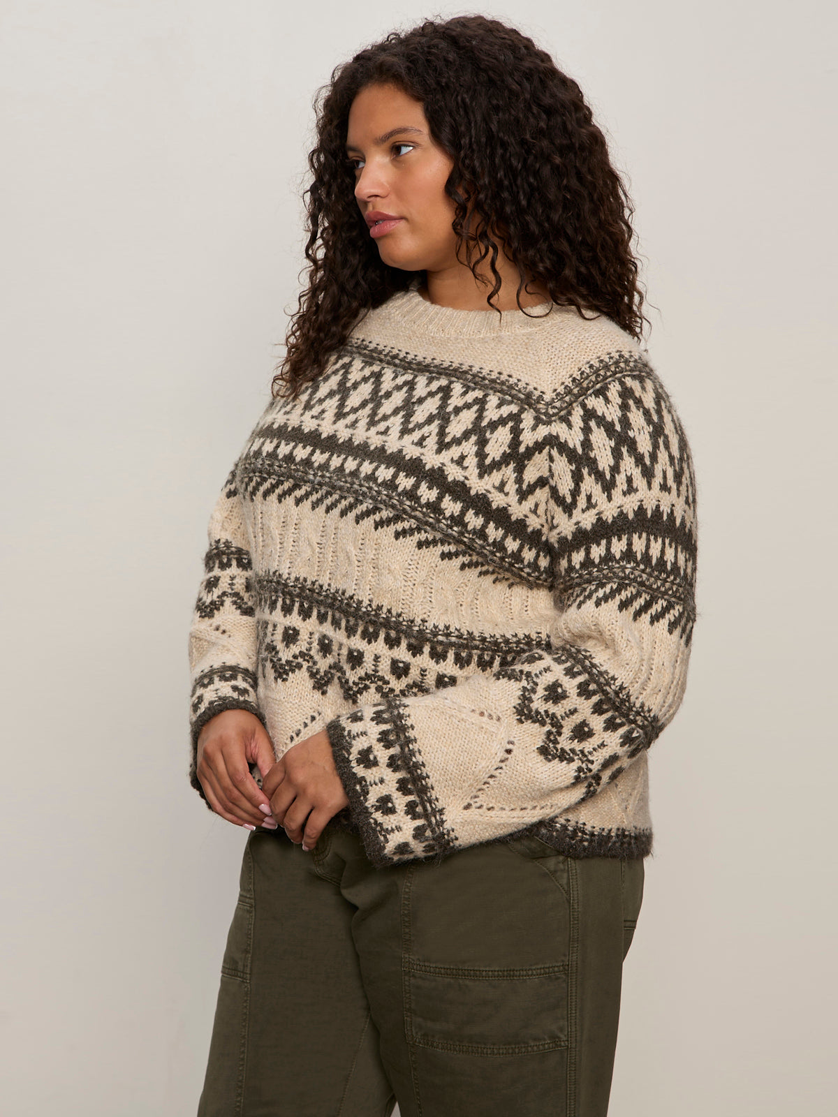 A woman with curly hair wears the Sanctuary Clothing fairisle sweater in light oat multi (extended sizing) and olive green pants, standing against a plain light background and looking to the side.