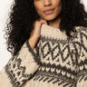 A woman with curly dark hair wears the Sanctuary Clothing fairisle crew neck sweater in light oat multi, looking thoughtfully to the side with her hand gently touching her neck.