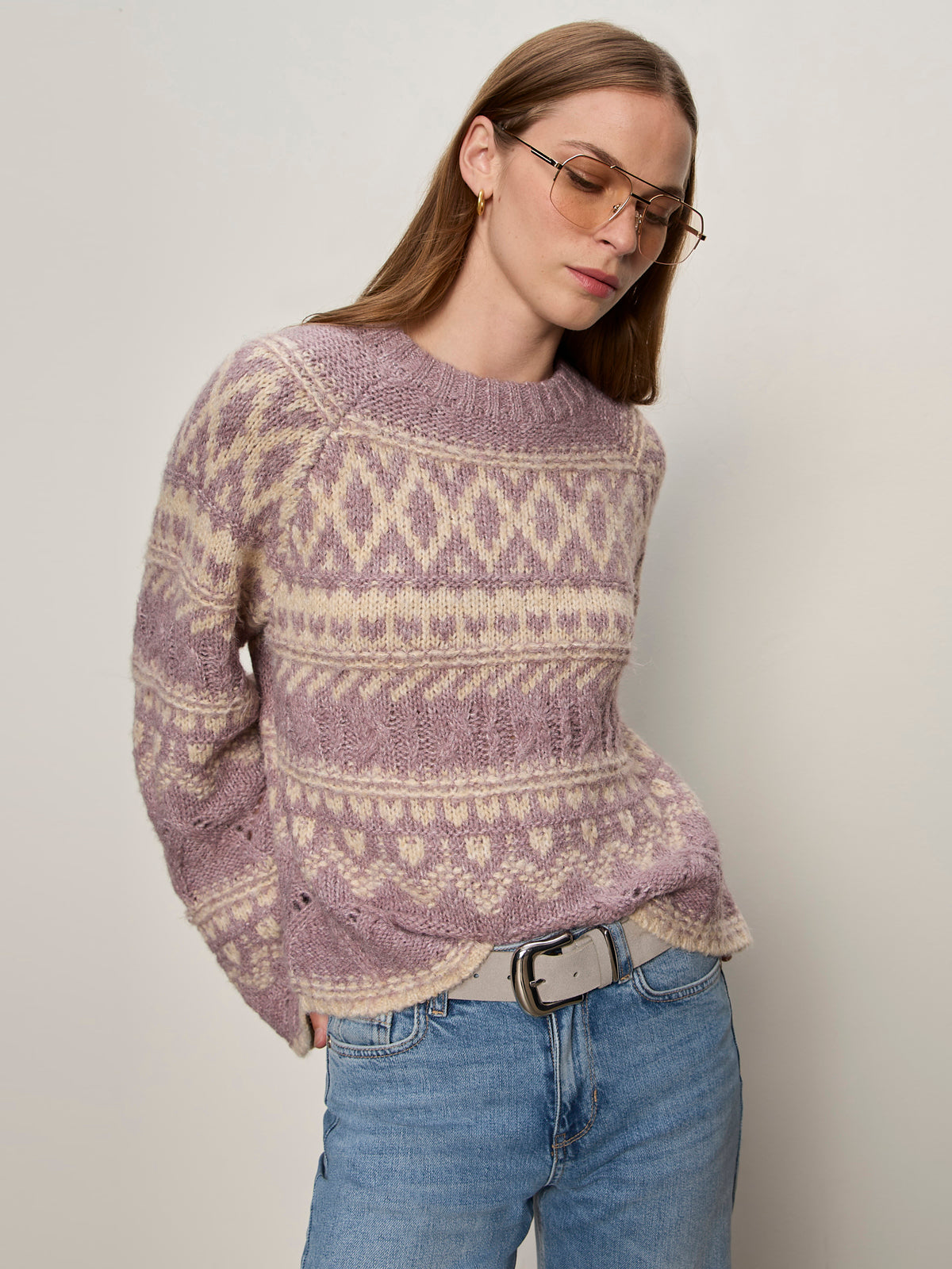 A person wearing the Sanctuary Clothing fairisle crew neck sweater in twilight mauve multi stands against a plain off-white background, paired with tinted sunglasses, light blue jeans, and a gray belt.